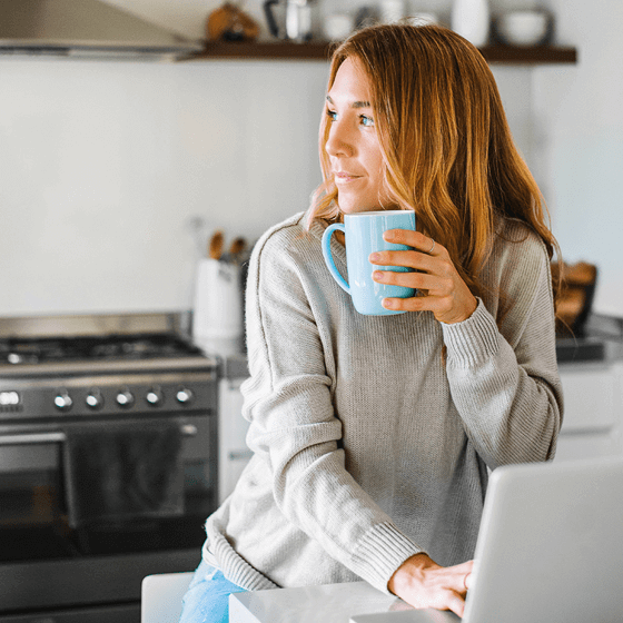 Kvinne på kjøkkenet ser ut av vinduet, holder kaffekrus og bruker bærbar PC