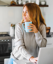 Kvinne på kjøkkenet ser ut av vinduet, holder kaffekrus og bruker bærbar PC